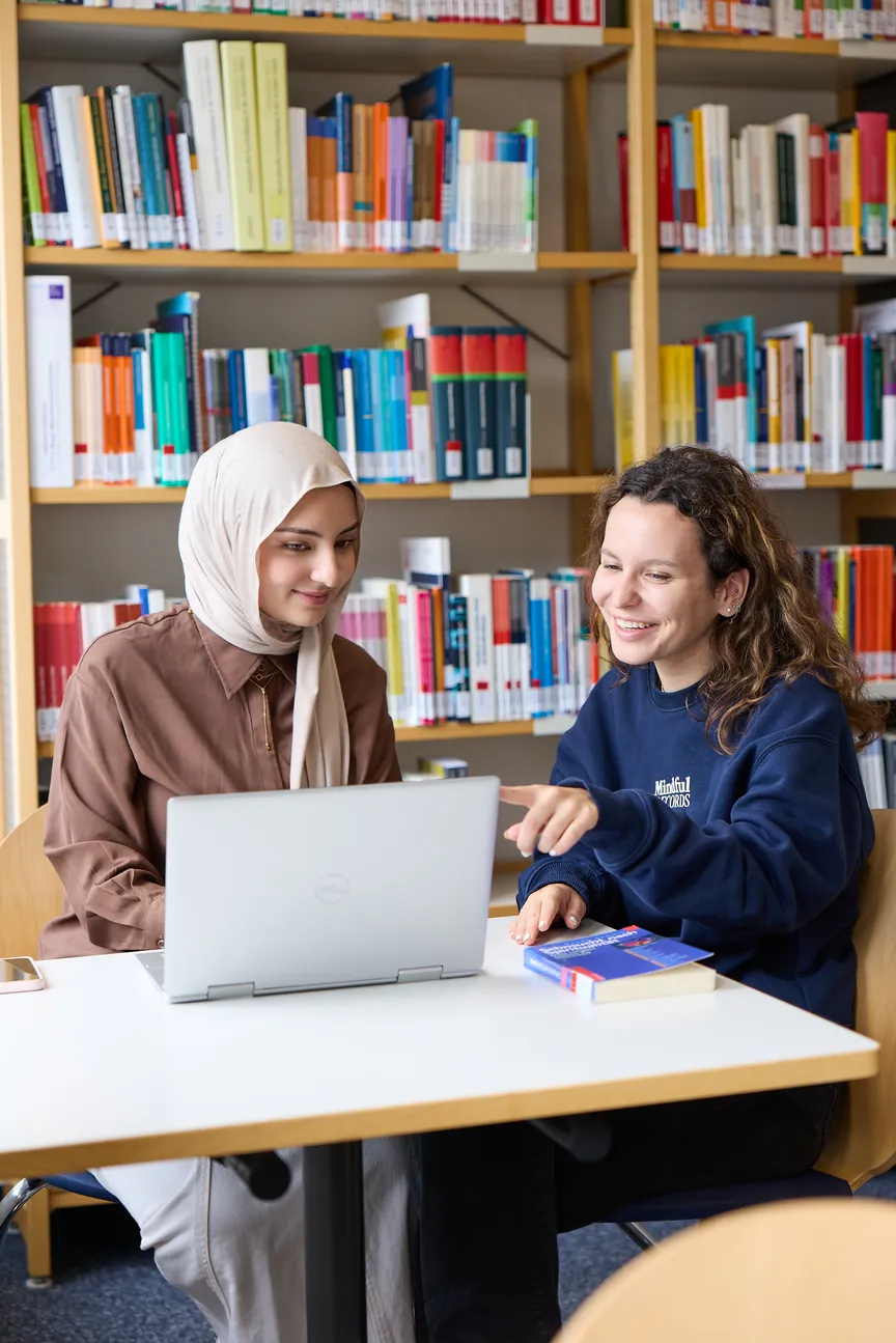 Zwei weibliche Studierende sitzen an Arbeitsplätzen in einer Bibliothek und reden miteinander