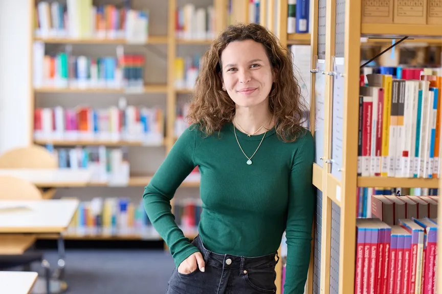 Eine Studierende steht an einem Bücherregal und lächelt