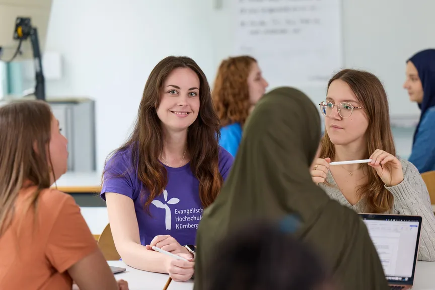 Eine Studierende sitzt in einer Gruppe Studierender in einem Seminar und lächelt in die Kamera
