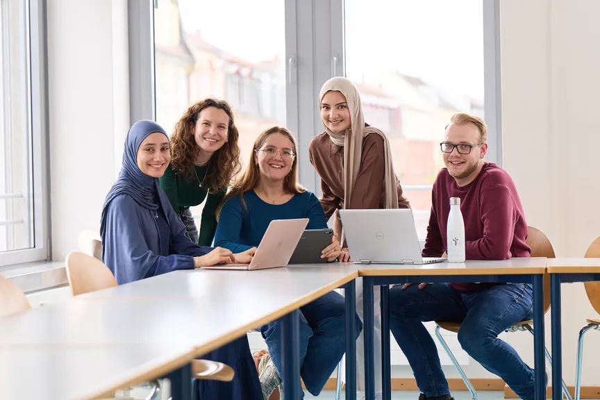 Eine Gruppe Studierender arbeitet an Laptops und lächelt in die Kamera