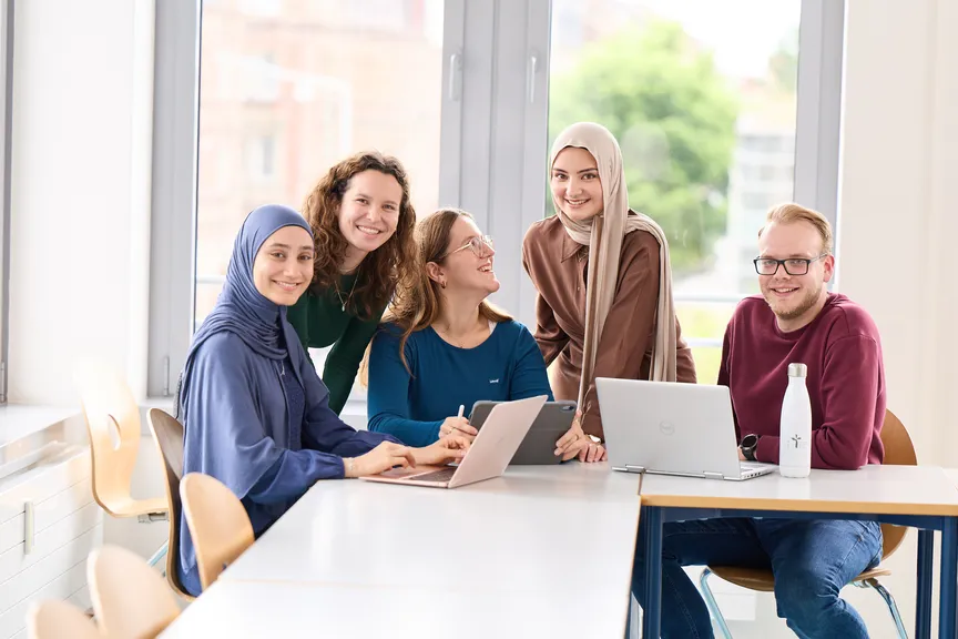 Eine Gruppe Studierender arbeitet an Laptops und lächelt in die Kamera
